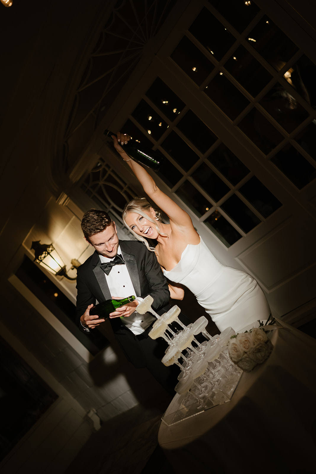 Bride cheers and raises a bottle while the groom pours champagne over their tiered glass tower at night.